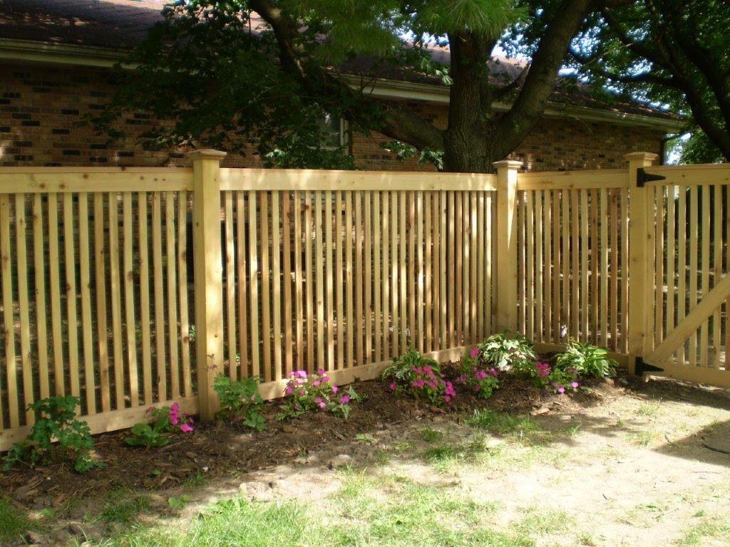 Wooden picket fence with gate, flowers in front, brick house and tree in background.