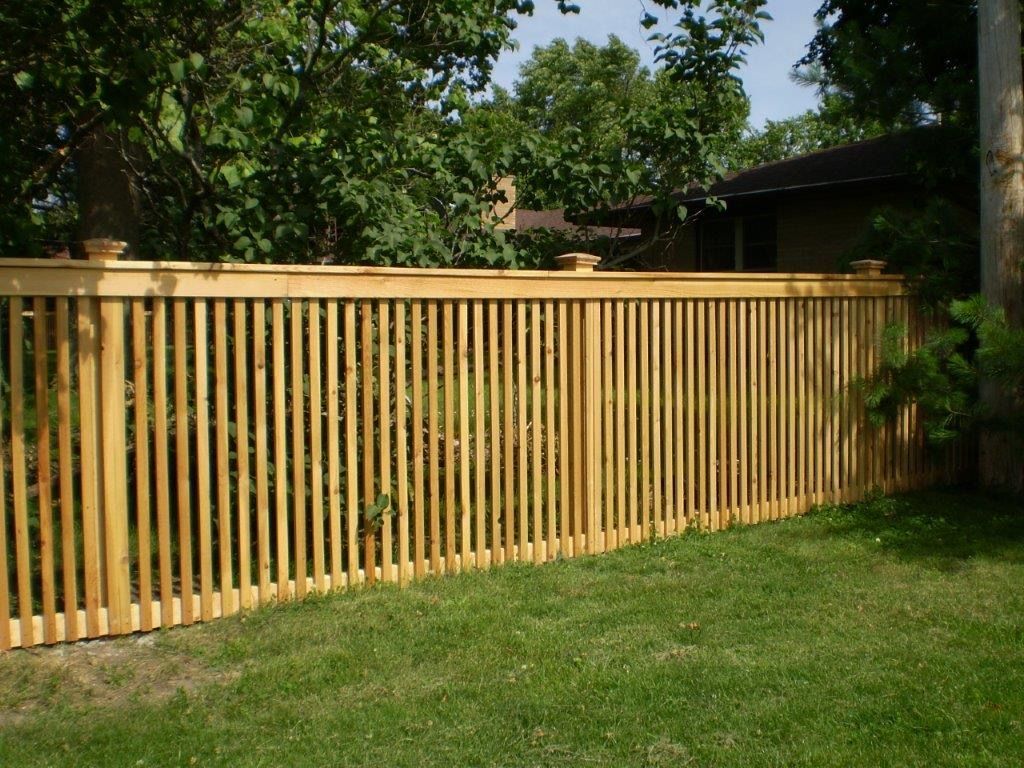 Wooden slatted fence in a grassy yard, partially obscuring a house and trees.