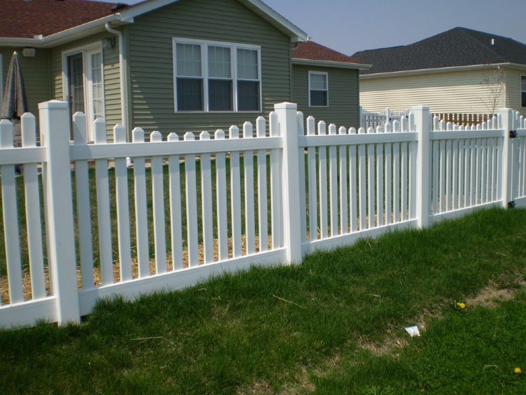 White picket fence in front of a green-sided house and green lawn.