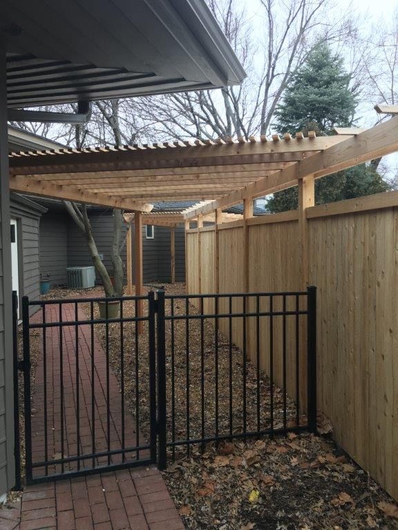 Pergola extending over a brick path, framed by a black metal fence and wooden fence.