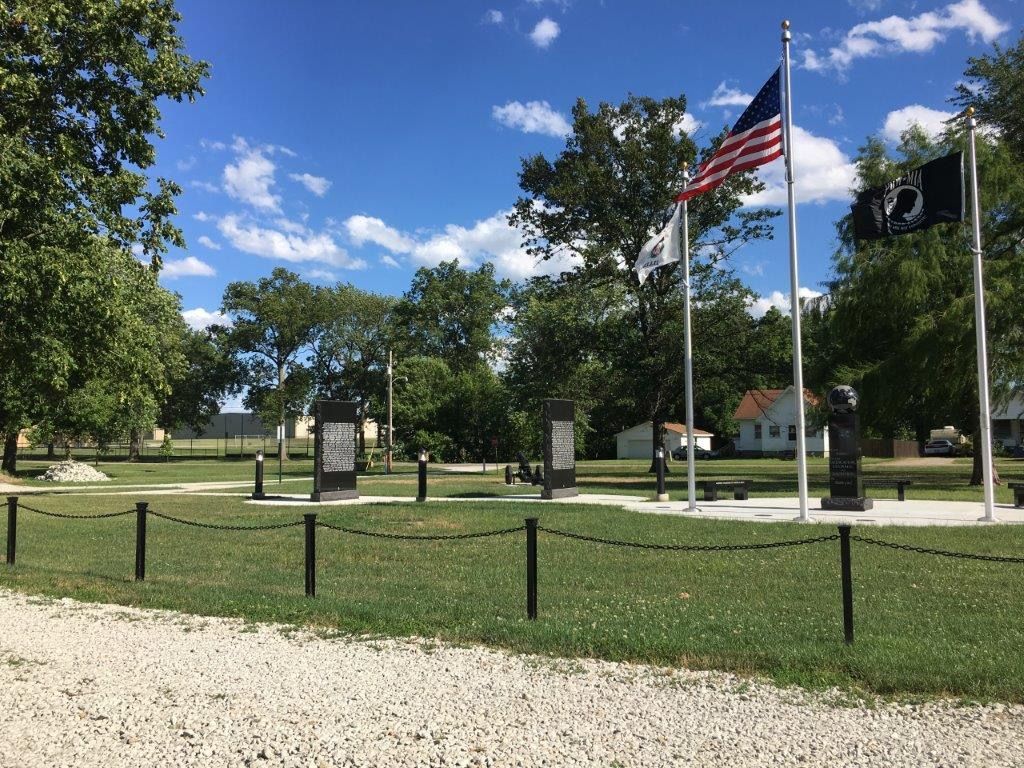 War memorial with flags, monuments, and a statue on a grassy lawn under a blue sky.
