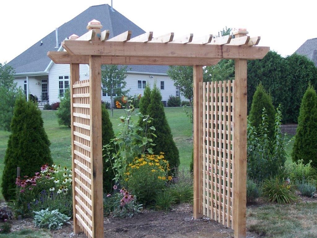 Wooden garden arbor with latticework side panels, surrounded by landscaping and a house in the background.