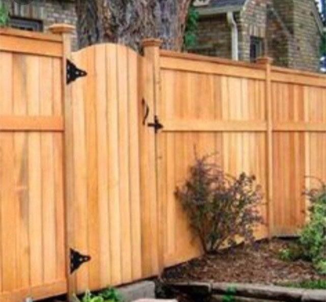 Wooden fence with gate, featuring black hinges and latch; plants in the foreground, brick house in the background.