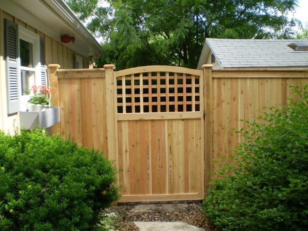 Wooden gate with lattice detail in a cedar fence, surrounded by green shrubs and a home.