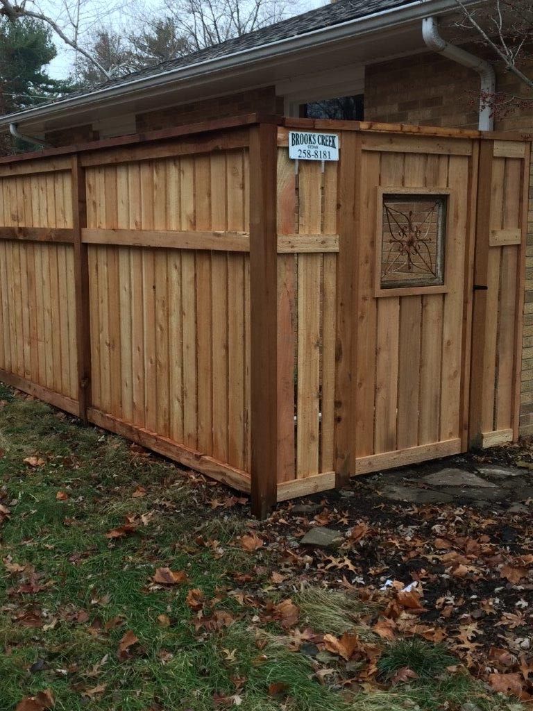 Wooden fence with gate, light brown boards, against a house. A decorative window is on the gate.