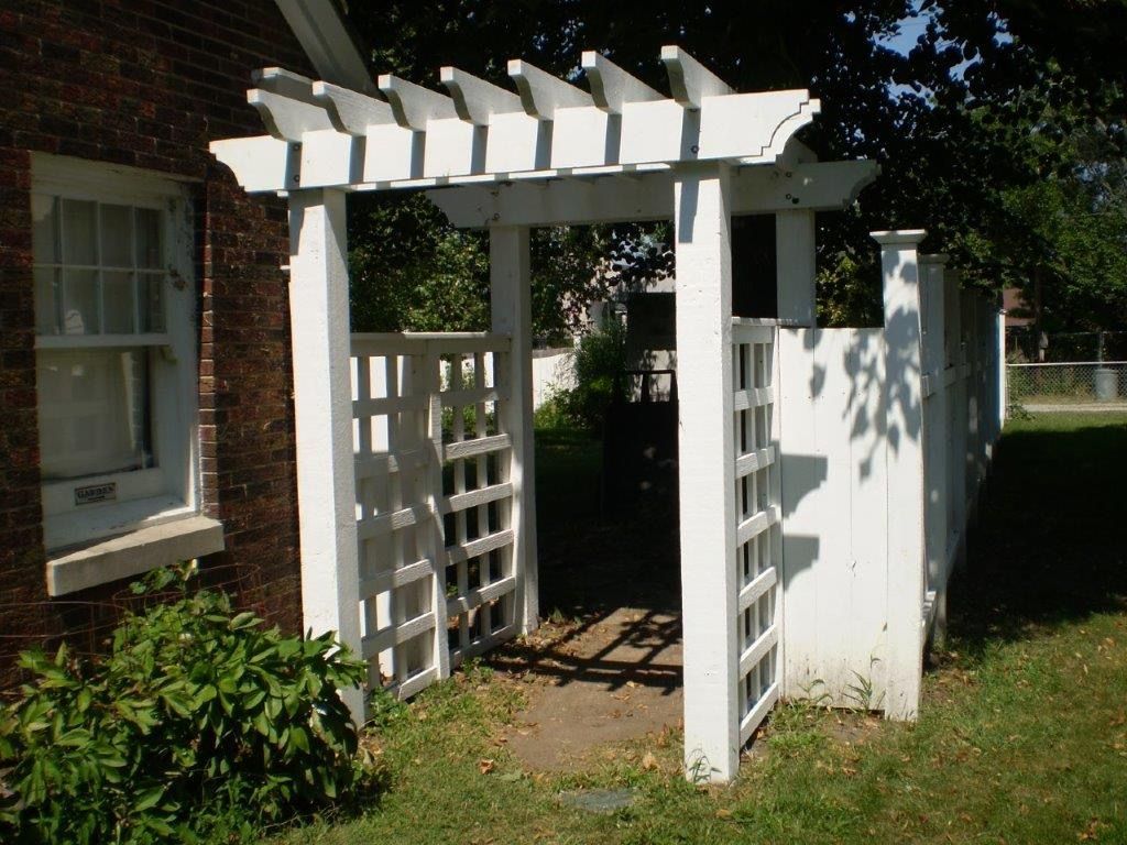 White pergola over a latticed gate and fence, next to a brick building.