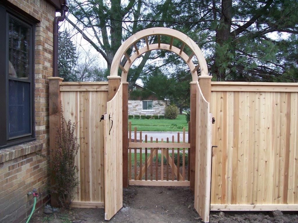 Wooden archway gate in a cedar fence, leading to a small picket fence and yard.