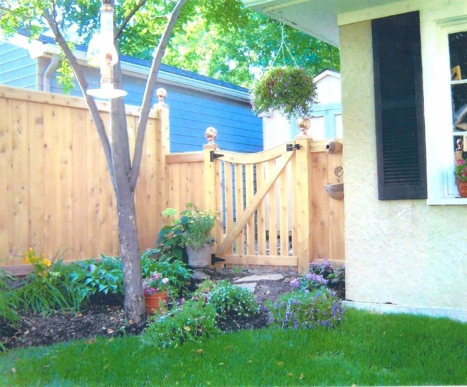 Wooden fence and gate enclose a garden with flowers and tree; house with shutter is on the right.