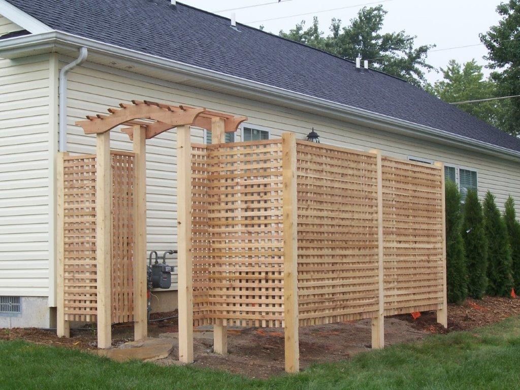 Wooden lattice outdoor shower structure against a house with a pergola-like top.