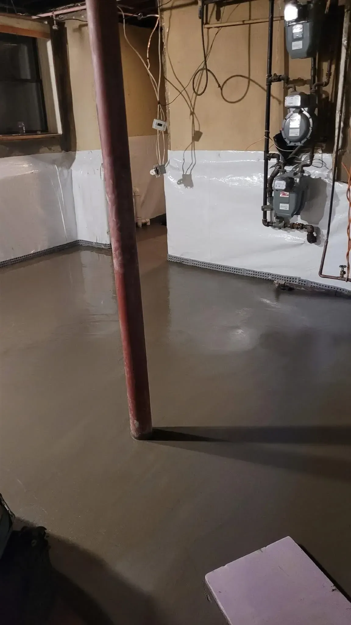 Basement with newly-laid gray flooring. A support pole stands in the center. White walls and utility equipment are visible.