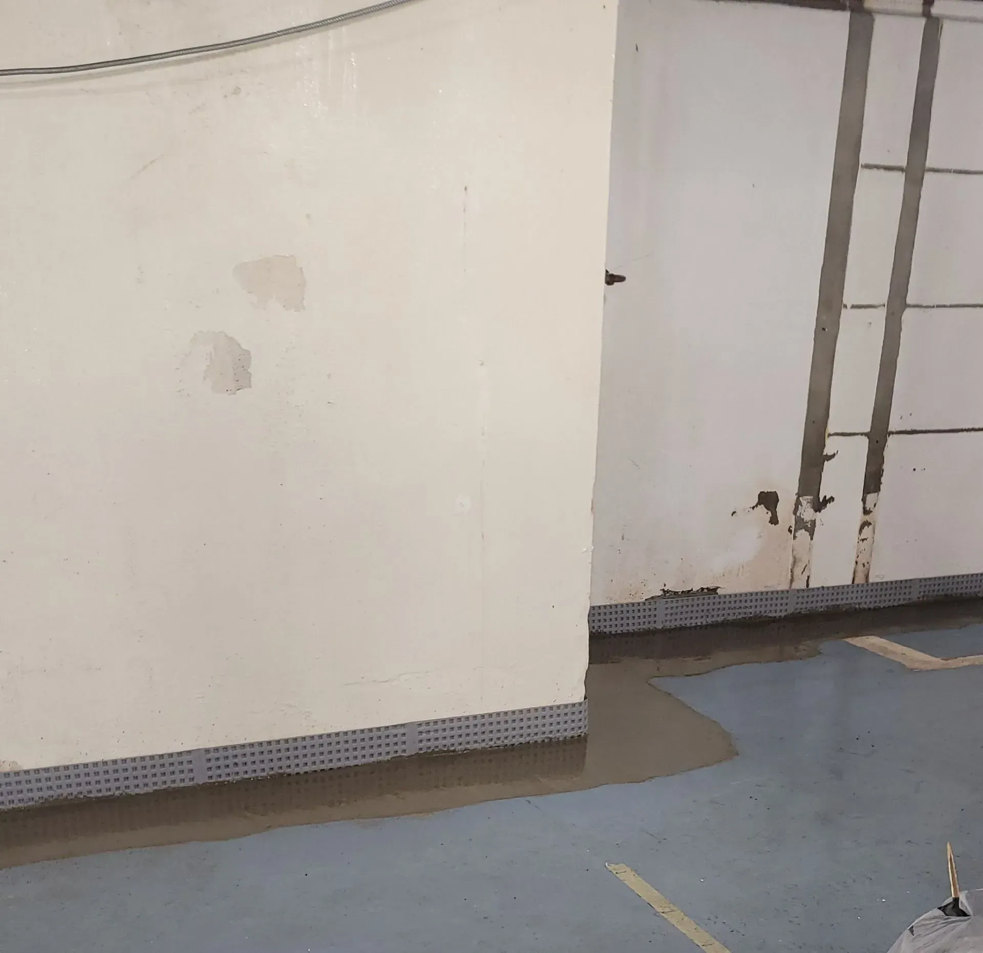 Gray, wet basement floor with water pooling near a wall with a plastic drainage system along the base.