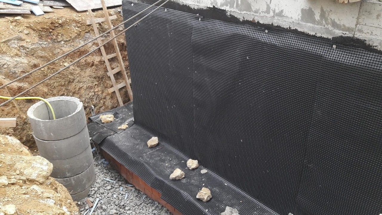 Black drainage membrane applied to a concrete foundation wall in an excavated area.