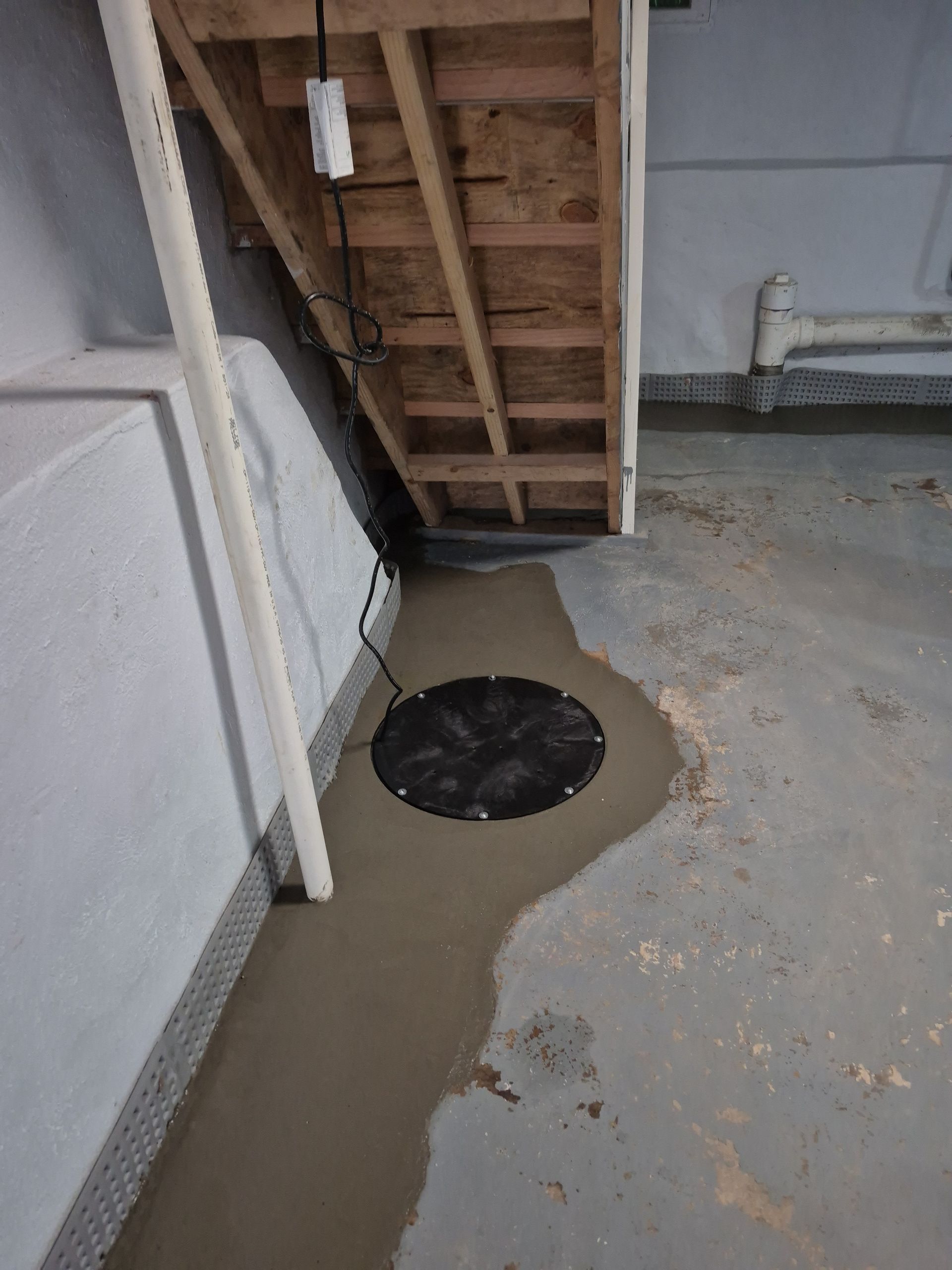 Basement floor with a sump pump and a new concrete patch under stairs and near a wall with white piping.
