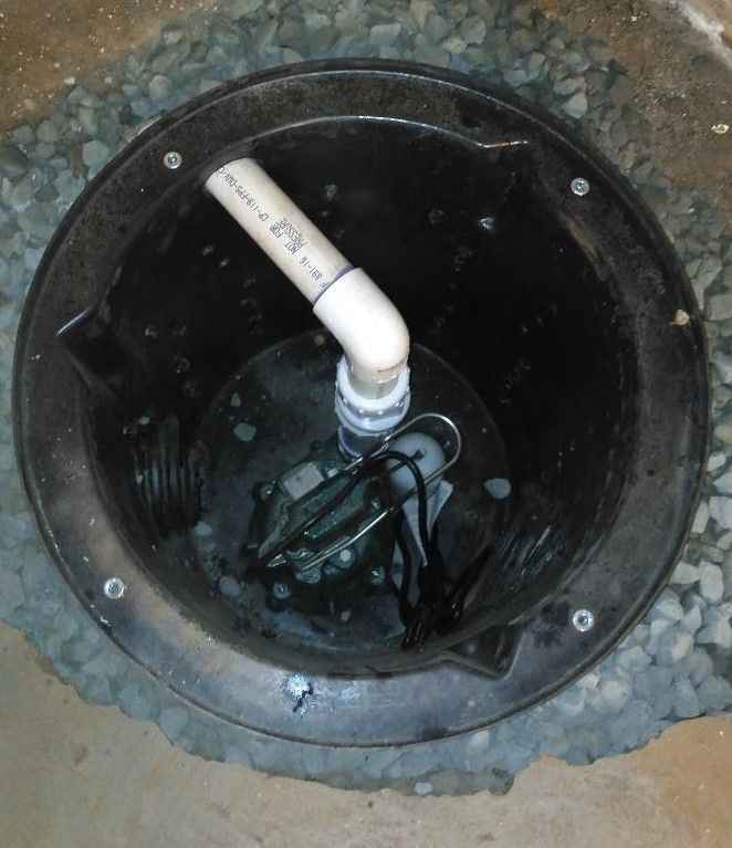 Black sump pump in a circular pit, with PVC pipe and surrounding gravel.