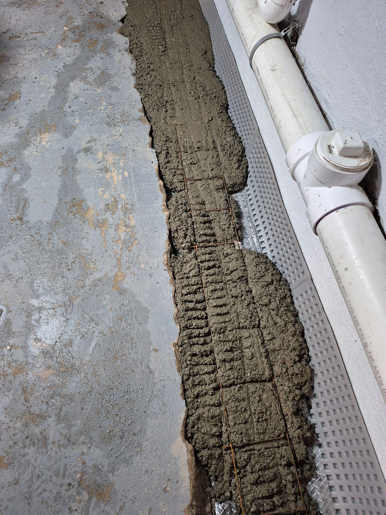 Concrete trench along a wall with drainage pipe and plastic mesh, likely for water management.