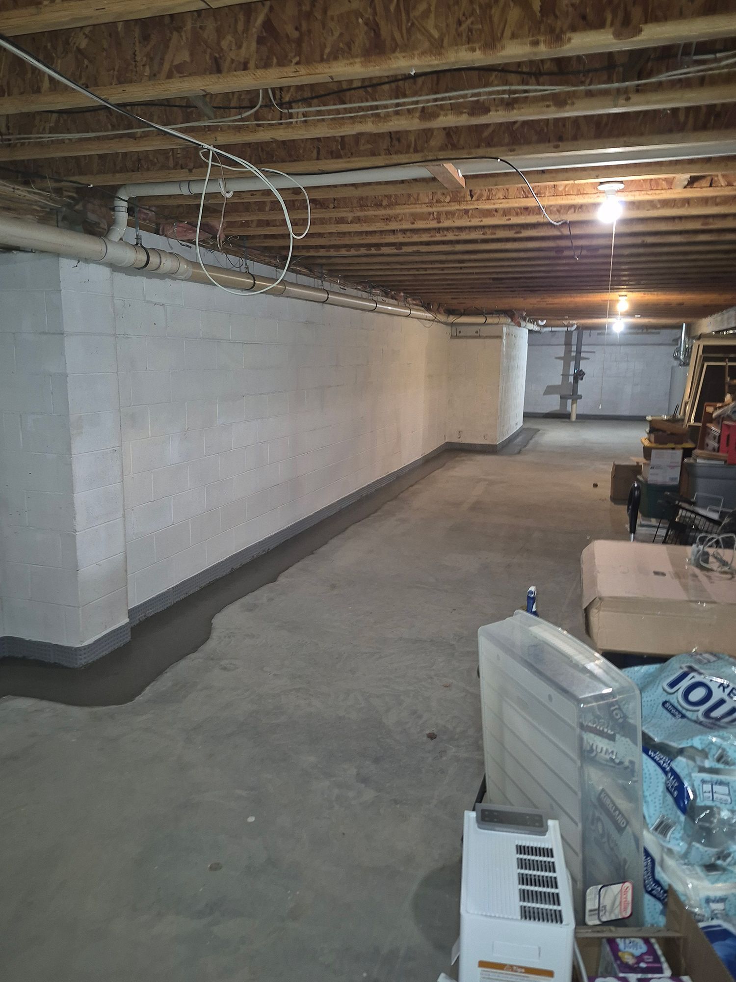 Basement with concrete walls, floor. Dark gray sealant applied along base. Ceiling beams, plumbing visible.
