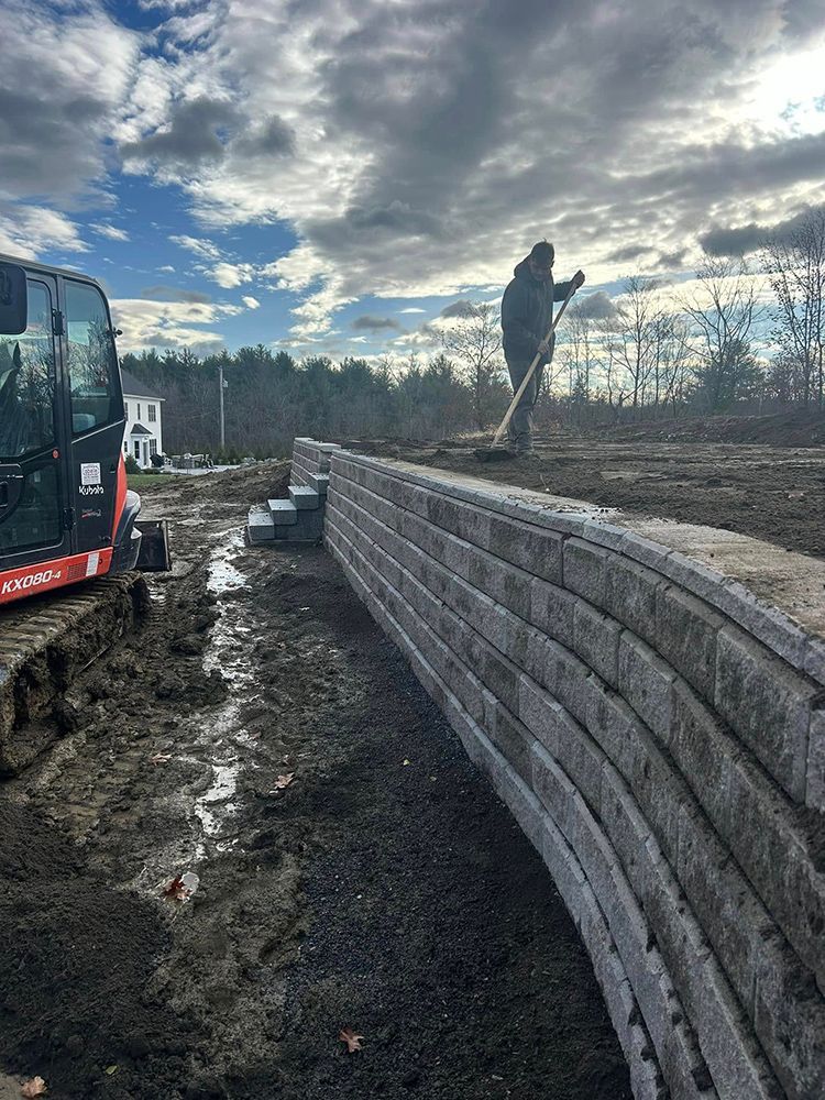 Construction site with a backhoe, retaining wall, and worker with a rake under a cloudy sky.