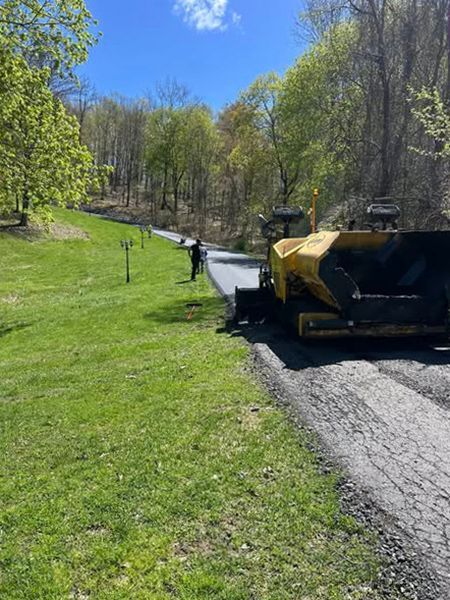 Road paving in a grassy, wooded area; asphalt paving machine and worker visible.