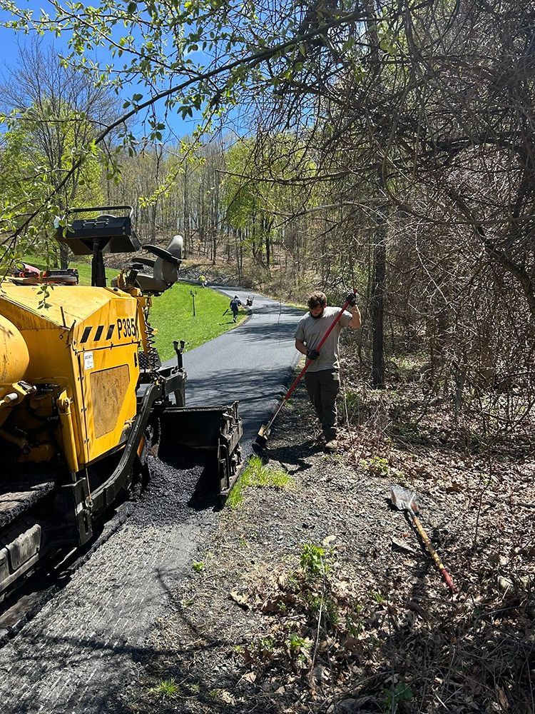Man paving a path with asphalt using a machine in a wooded area on a sunny day.
