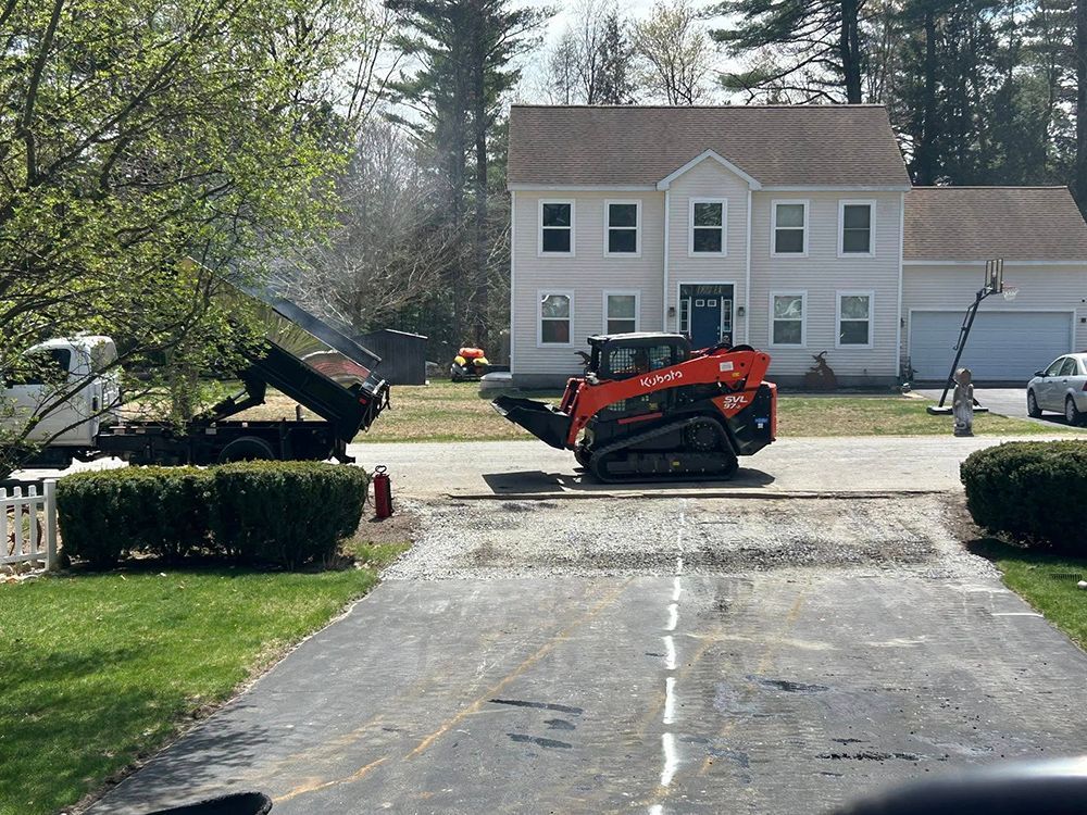 A red skid steer grinds up an asphalt driveway, with a dump truck and two-story house in the background.