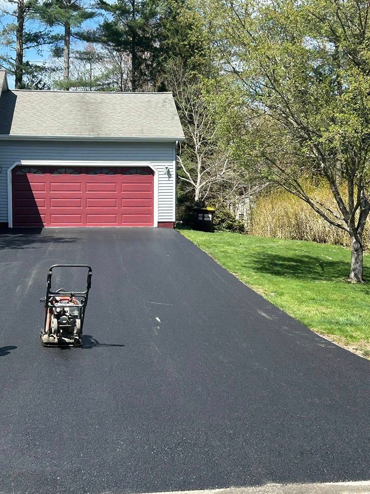 Newly paved black asphalt driveway in front of a red garage door and green lawn, with a compactor machine.