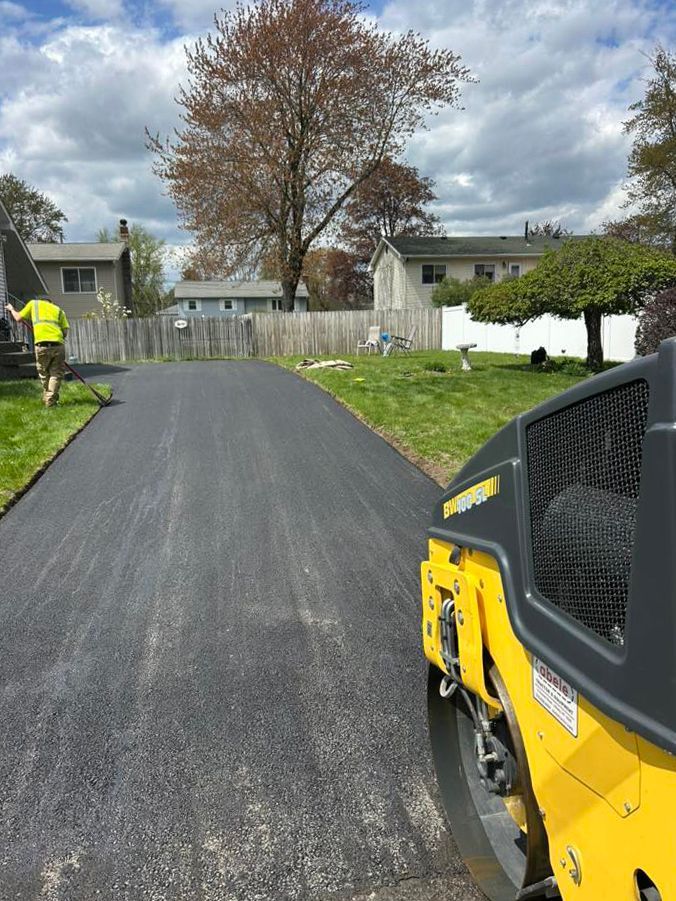Freshly paved driveway; worker raking asphalt, yellow roller in the foreground, houses and green grass in the background.