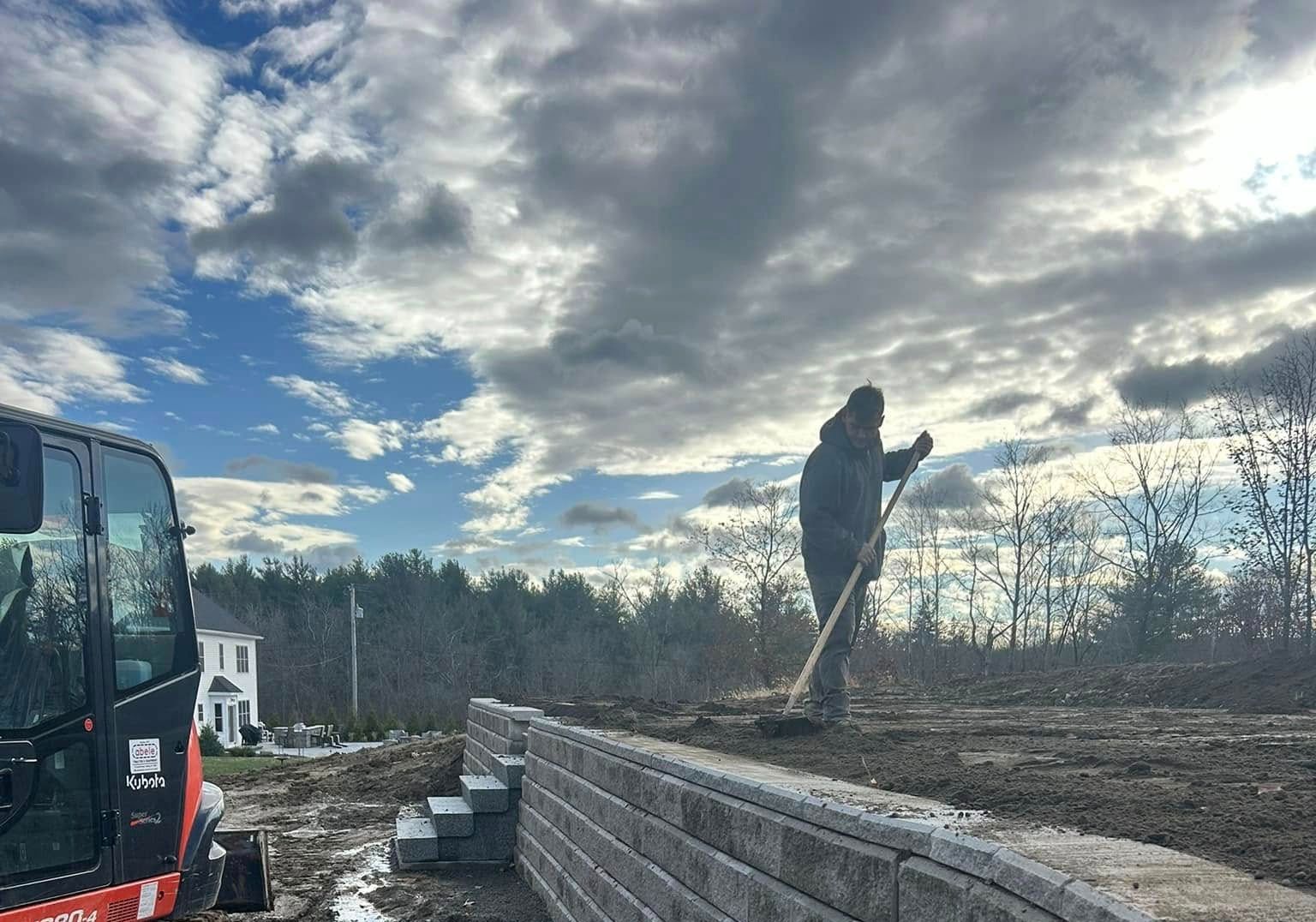 Person raking soil on a construction site with retaining wall, mini excavator, and cloudy sky background.