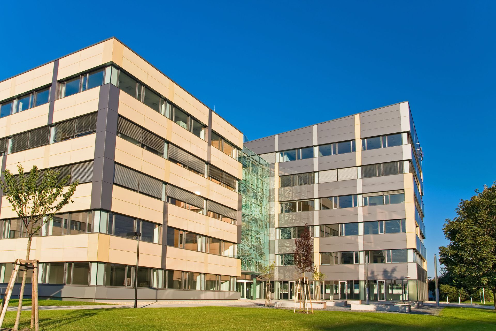 A modern office building with tan and grey panels, tall glass windows, and green landscaping against a clear blue sky.