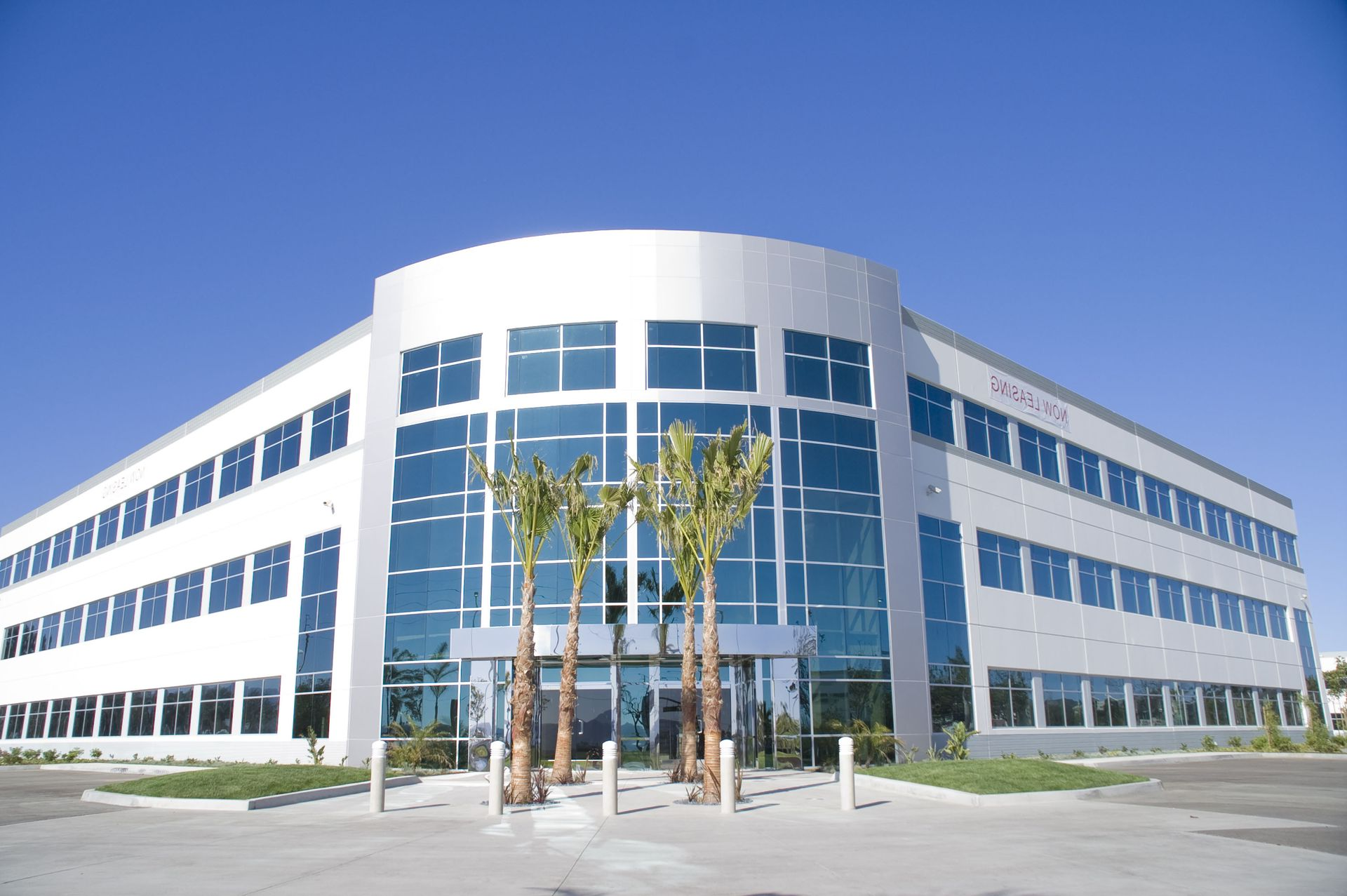Modern three-story office building with a curved glass facade and palm trees in front under a clear blue sky.