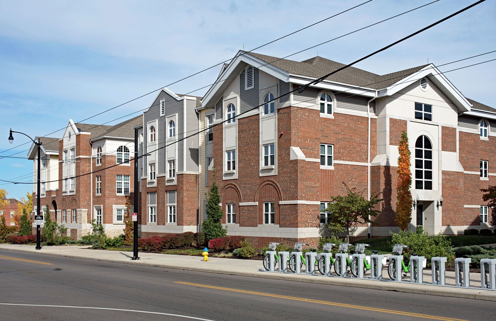 A four-story brick apartment building on a street with a bicycle rack along the sidewalk under a blue sky.