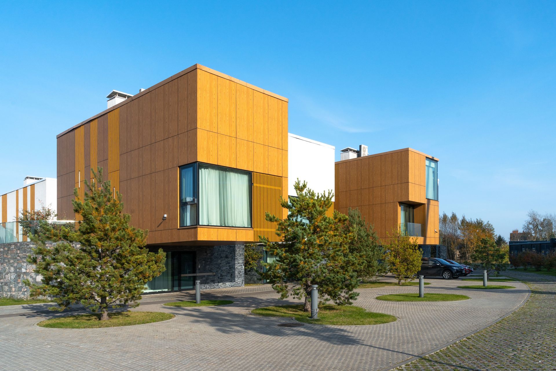Modern two-story homes with wood-paneled facades and large windows, situated on a stone-paved lot with small pine trees.