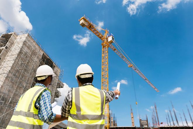 Two workers in hard hats and safety vests stand at a construction site, looking up at a crane against a blue sky.