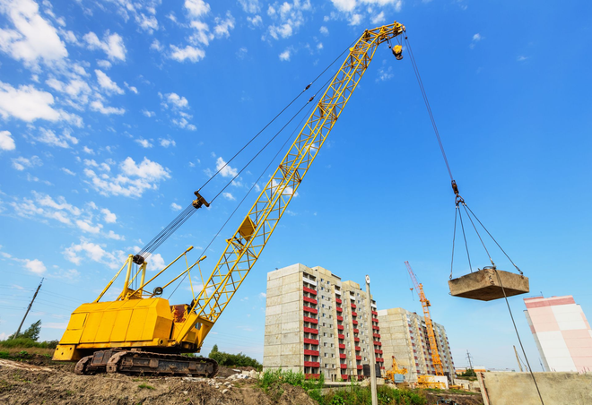 A large yellow crawler crane lifts a heavy concrete slab against a clear blue sky at a construction site.