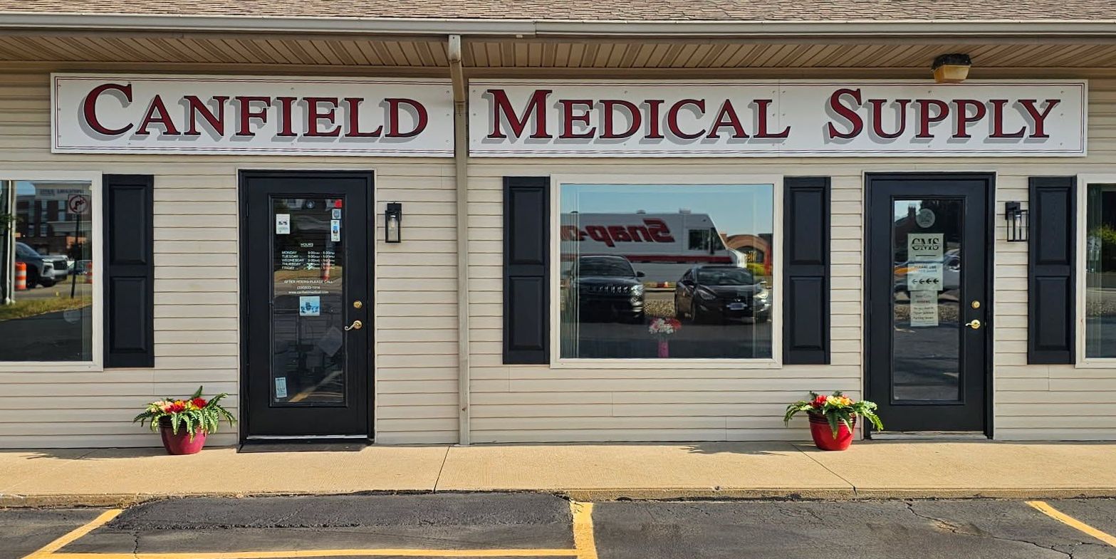 Canfield Medical Supply store front with beige siding, black doors and sign, and flowers in pots.