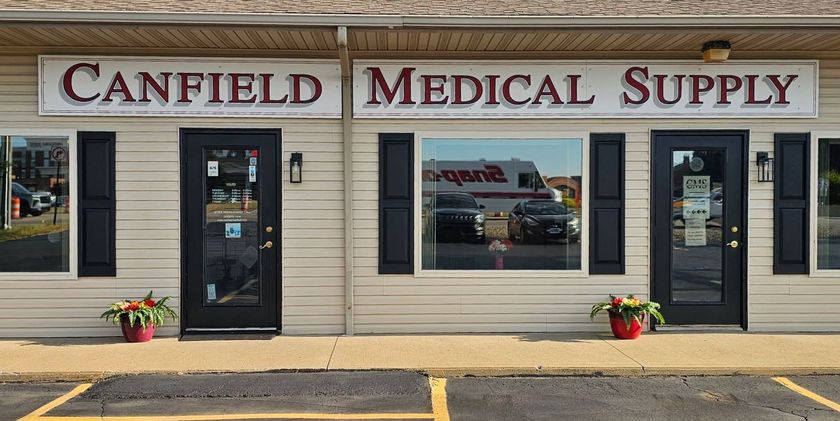Canfield Medical Supply store front with beige siding, black doors and sign, and flowers in pots.