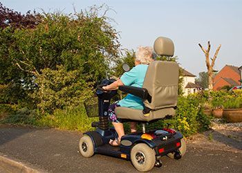 Person on mobility scooter, driving on a paved path next to a green hedge, small houses in the background.