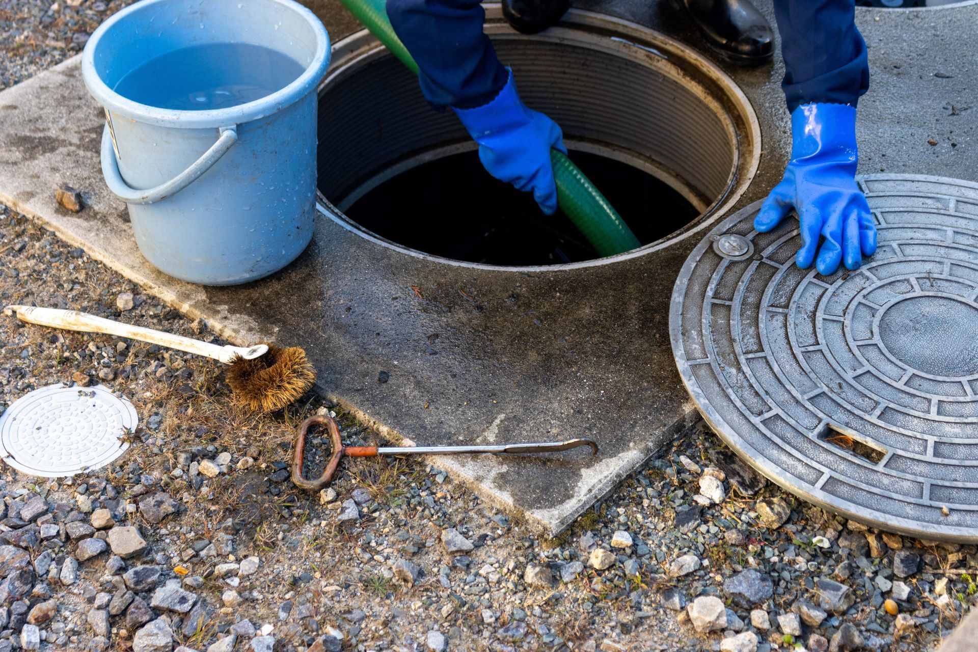 Worker opening a manhole beside a blue bucket and tools on gravel ground