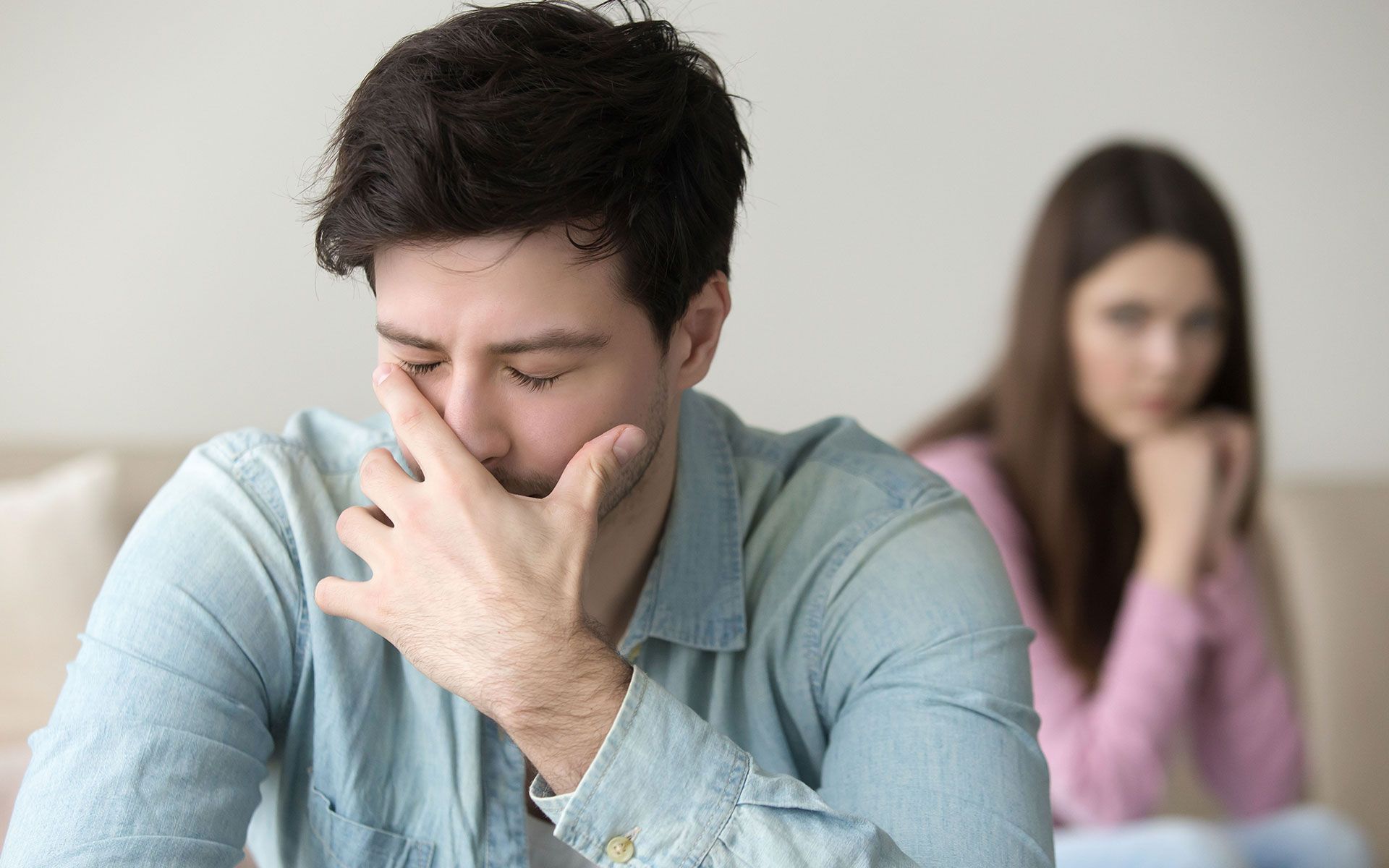 A man is covering his mouth with his hand while a woman sits behind him on a couch.