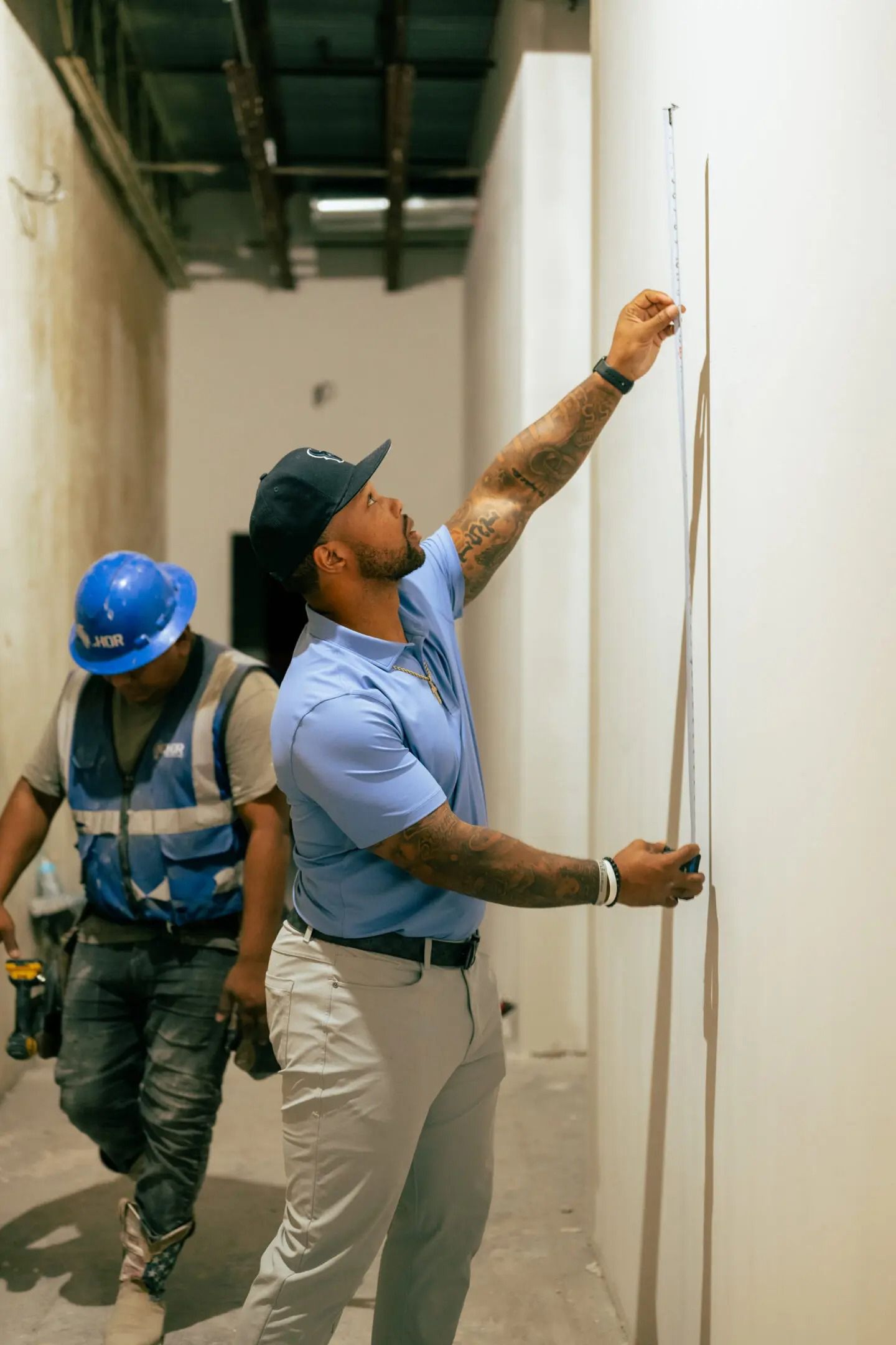 Two workers in hard hats install wall panels in a narrow hallway.