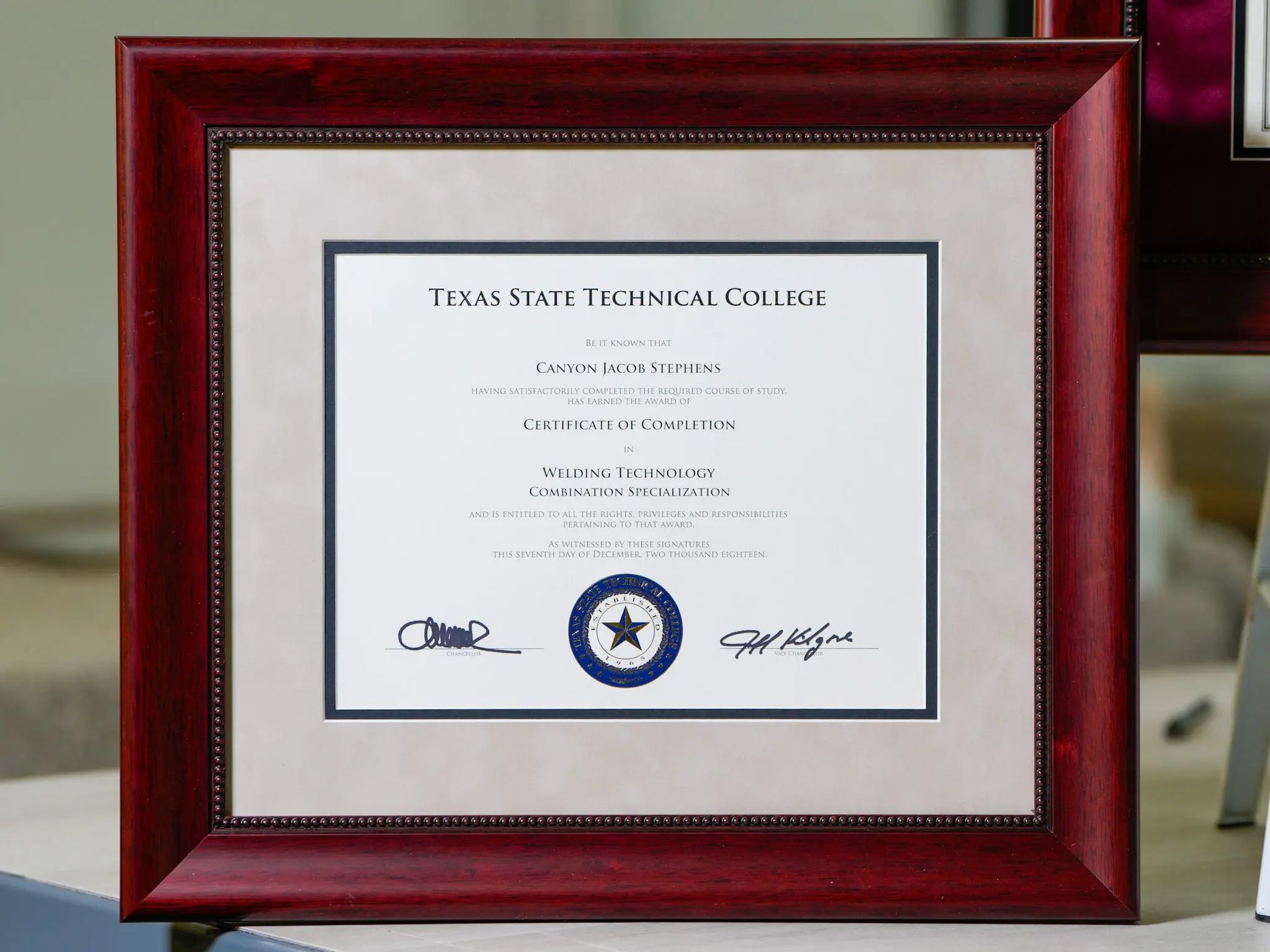 Framed Texas State Technical College diploma in a red wooden frame on a table