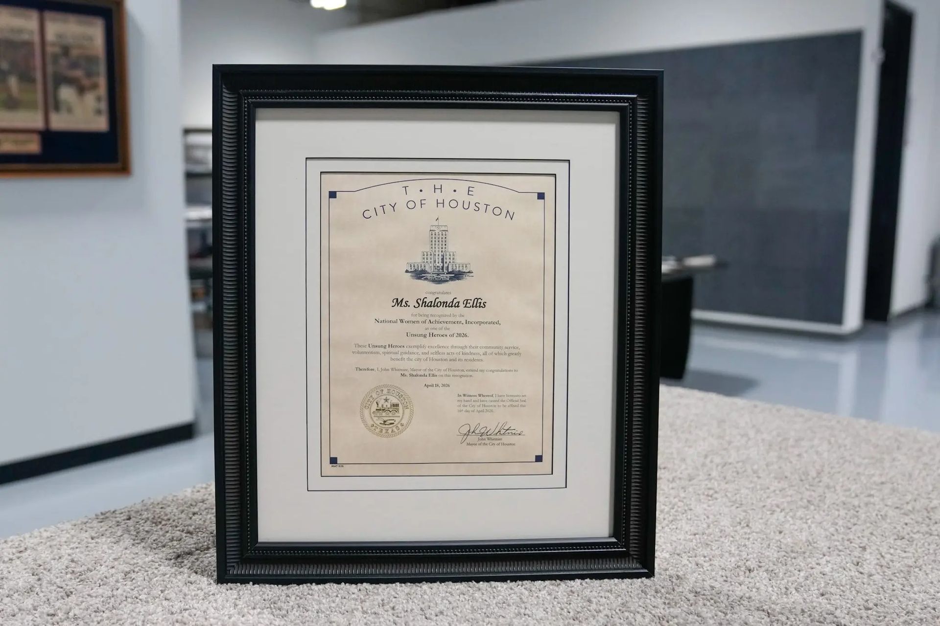 Framed certificate on a carpeted floor in a hallway, black frame with cream document.