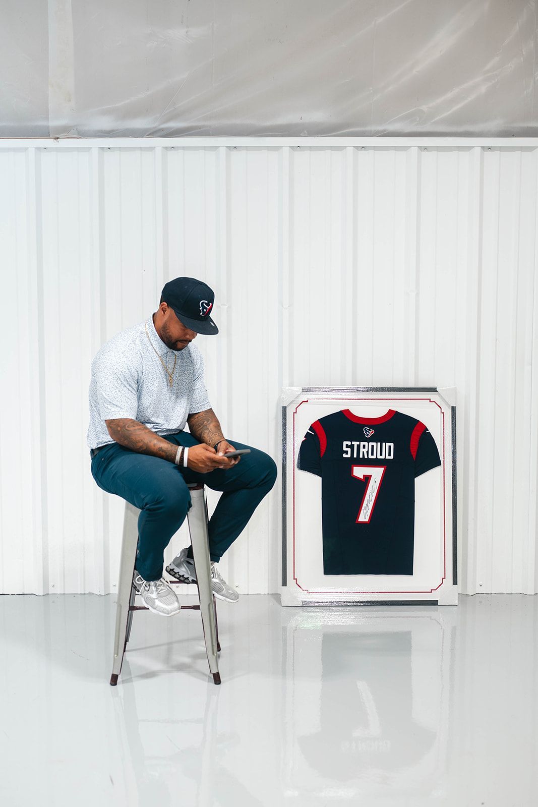 Man seated on a stool beside a framed black football jersey with red trim and number 7 in a white room