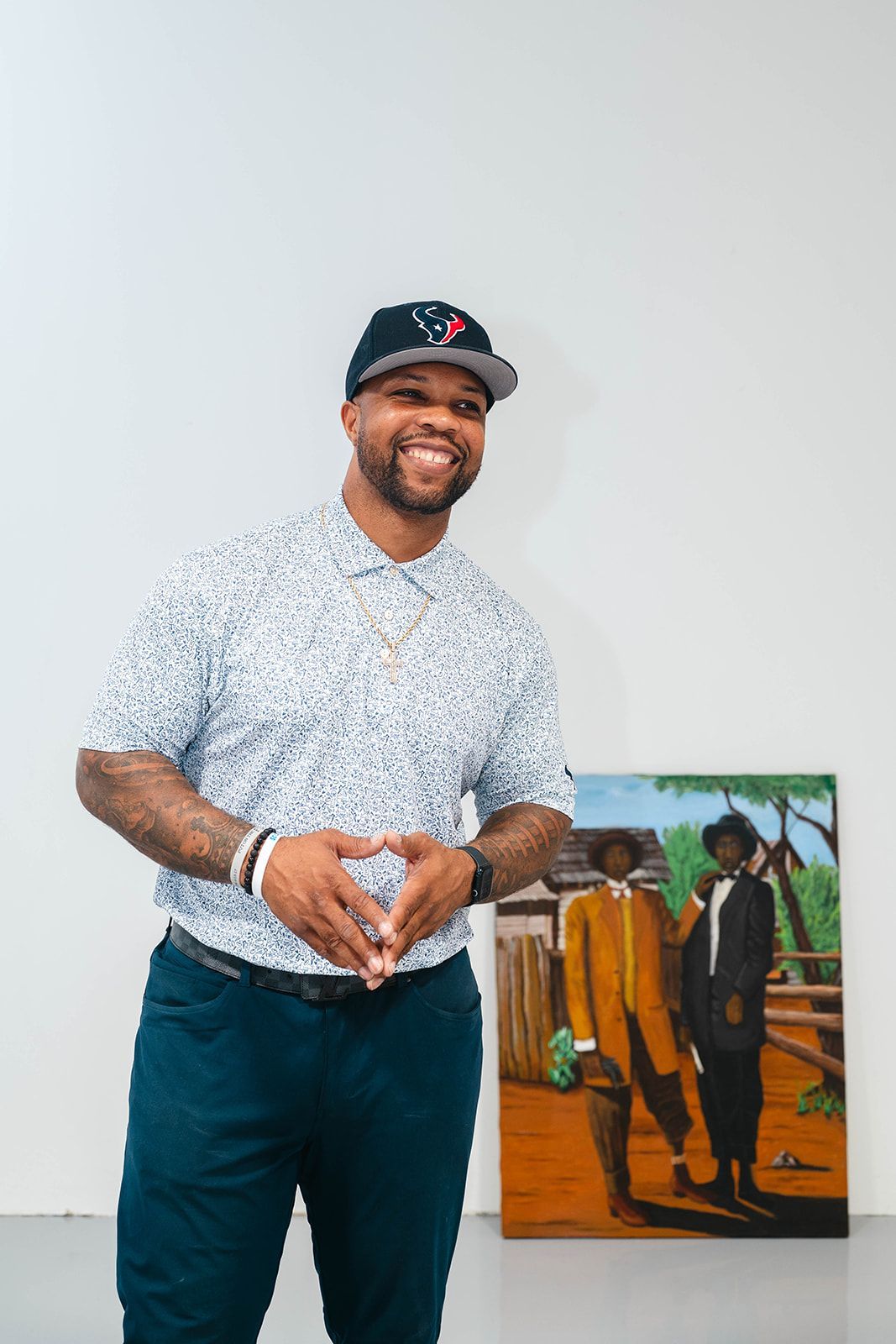 Man standing indoors beside a colorful painting, smiling and wearing a cap and patterned shirt.