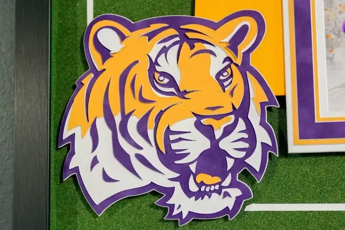 Tiger mascot logo on green turf, orange, purple, and white with an open mouth.