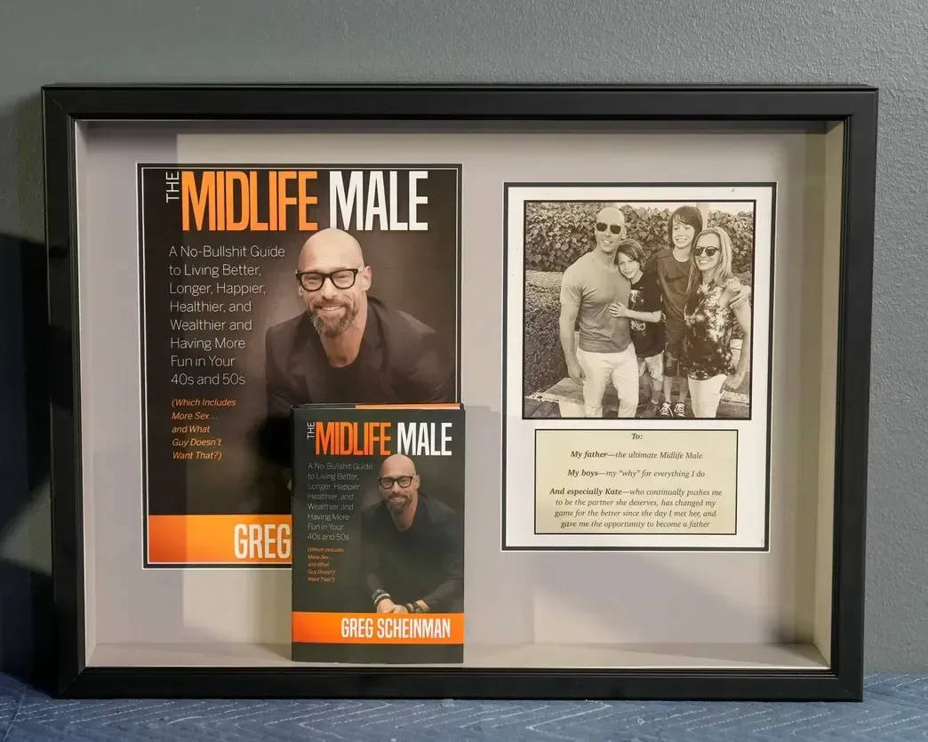 Framed display of two “MIDLIFE MALE” books beside a vintage sepia photo and note card.