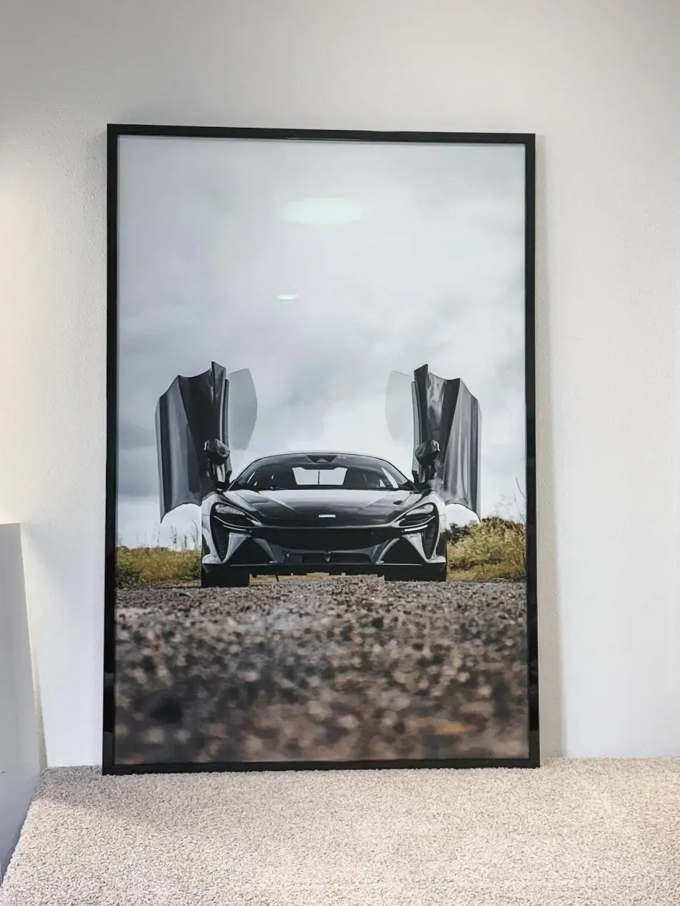 Framed poster of a black sports car on a gravel road with open doors, leaning against a wall.