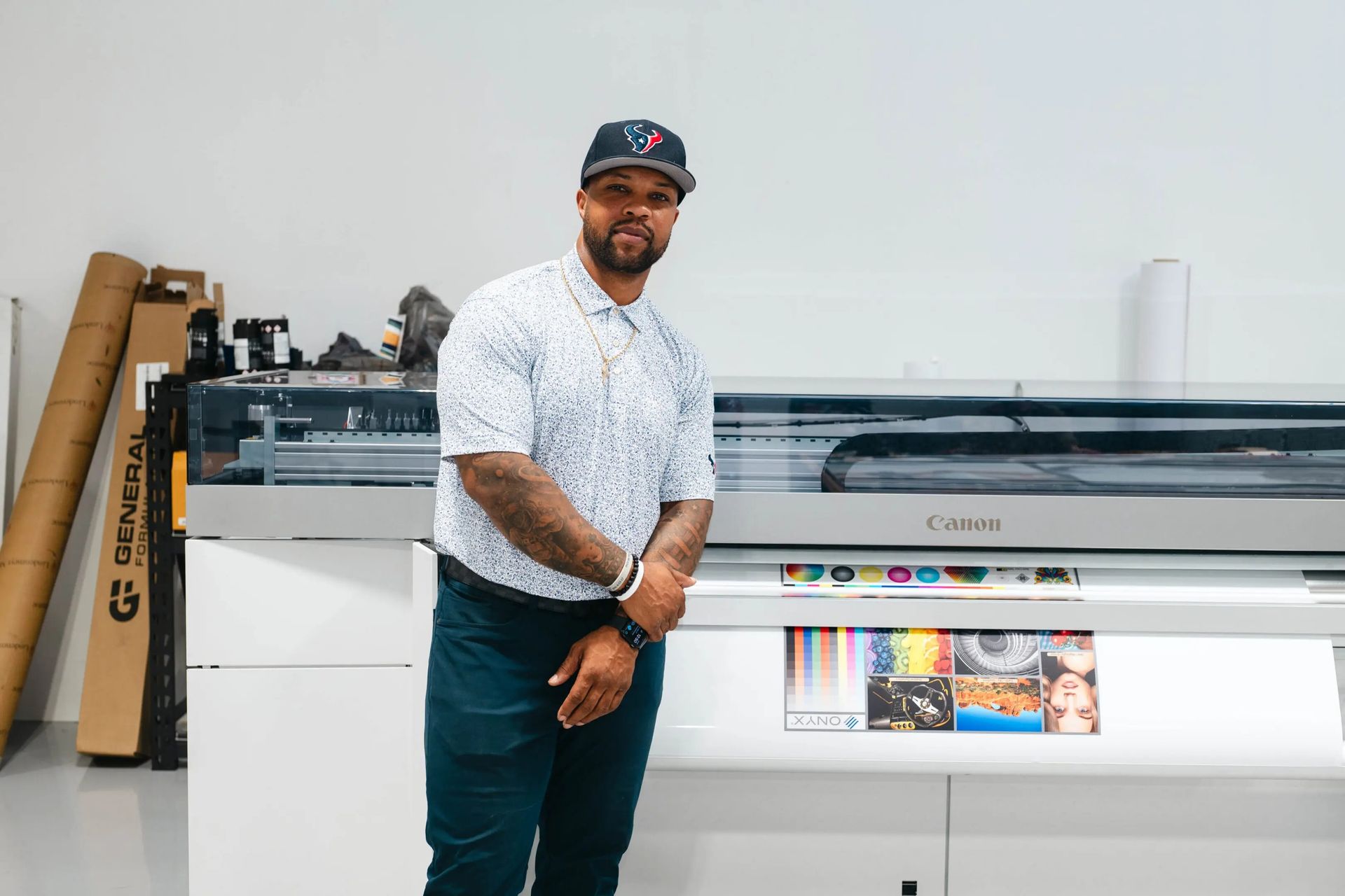Man standing beside a large-format printer in a print shop, with rolled posters and printed materials nearby