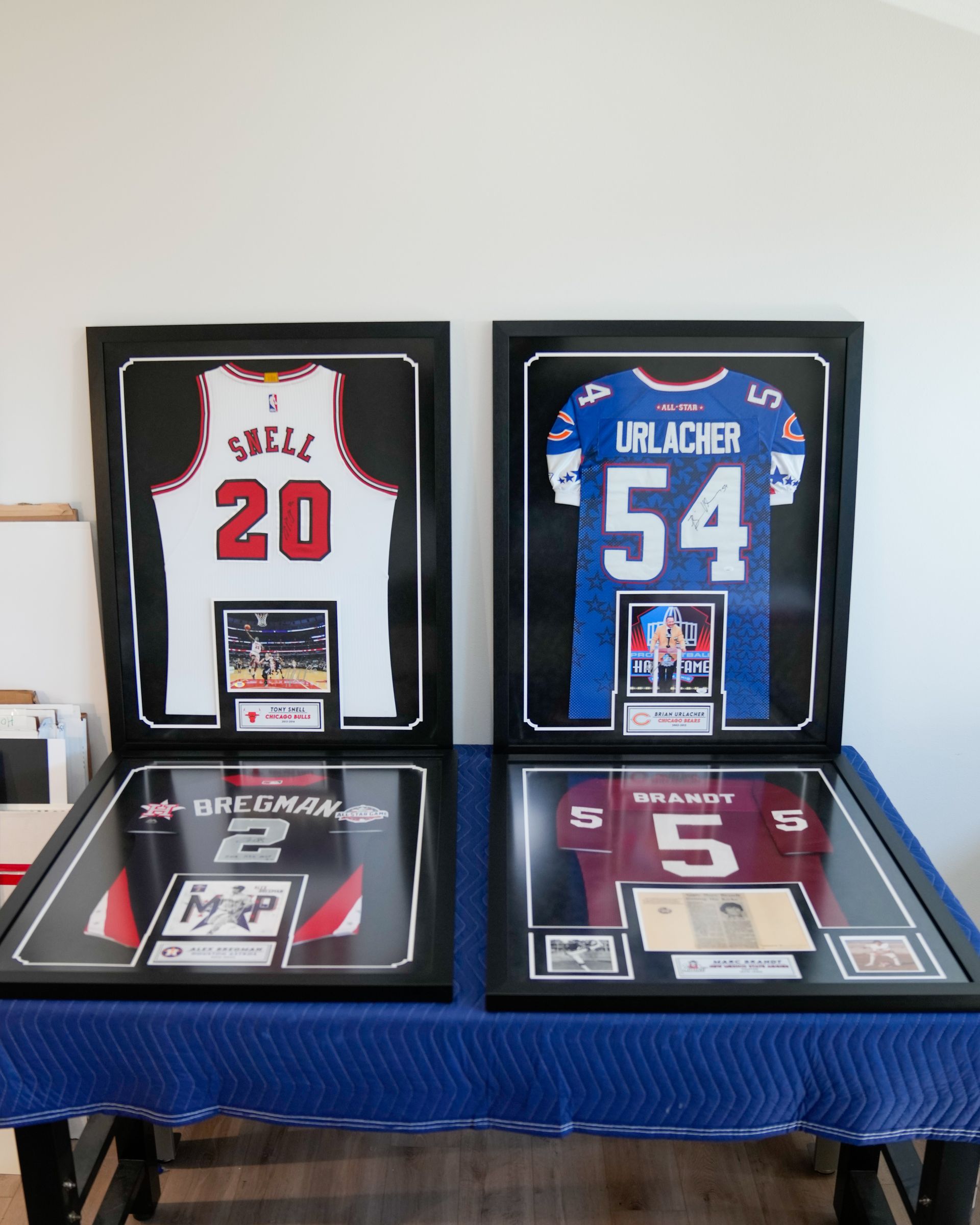 Framed sports jerseys and cards on a blue table, featuring white, blue, and maroon jerseys with numbers 20, 54, 2, and 5