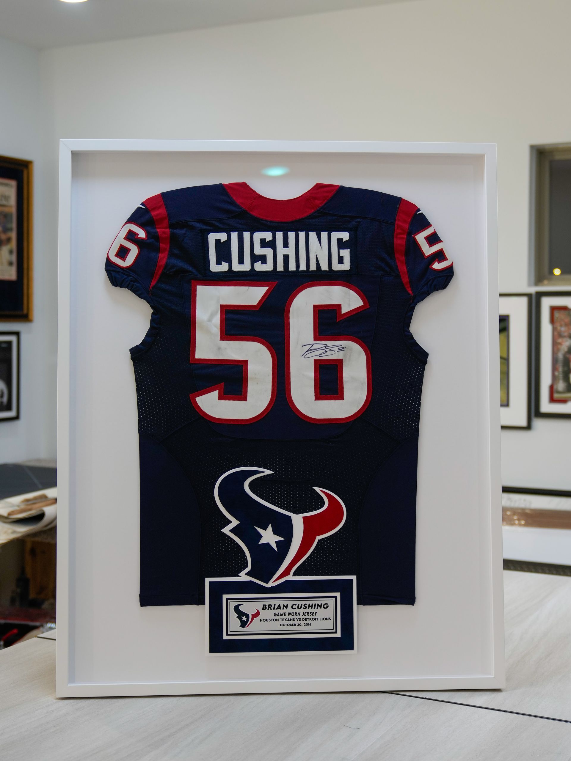 Framed Houston Texans jersey with “CUSHING 56” and team logo on a white wall