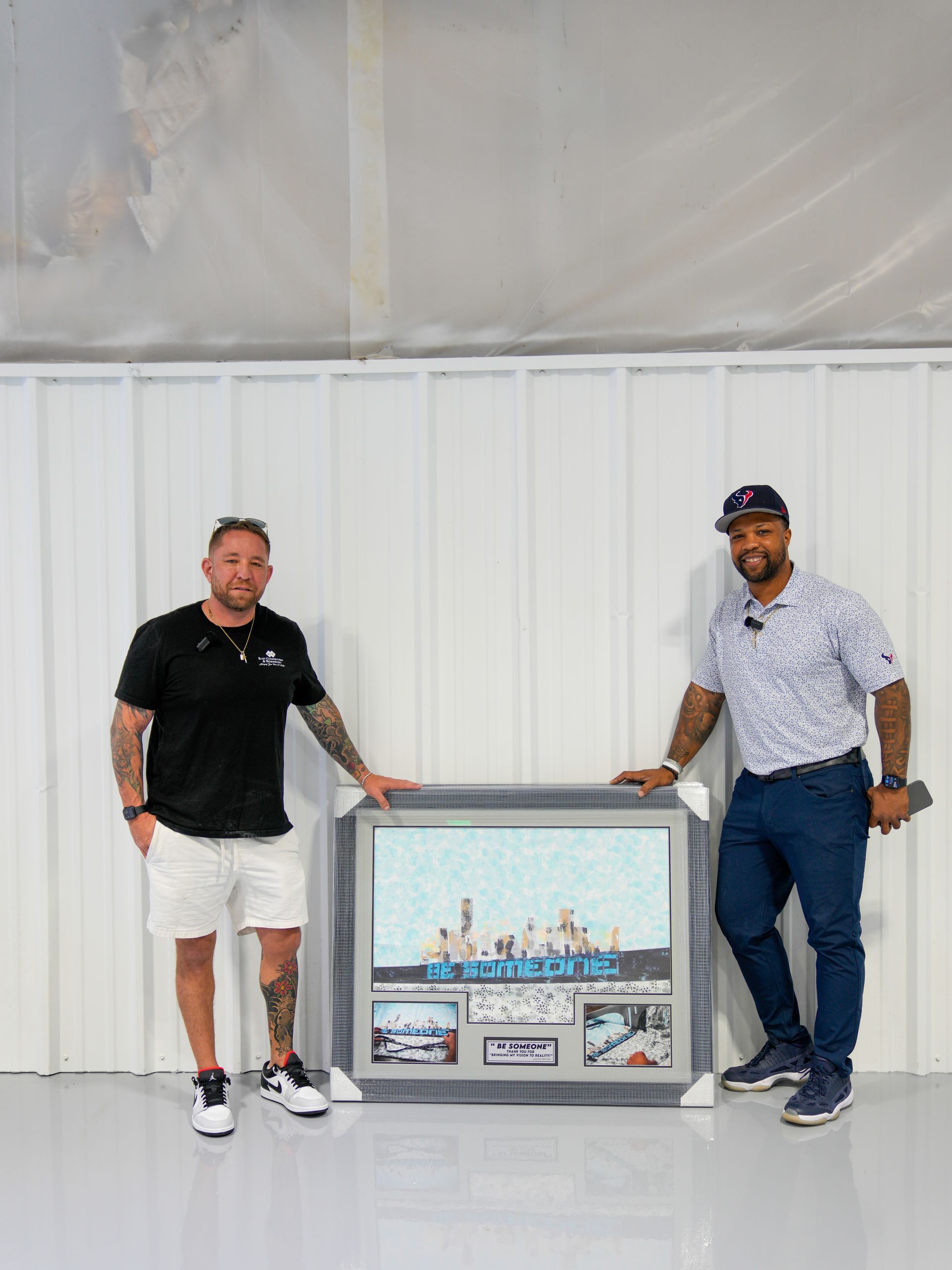 Two men stand beside a framed artwork leaning against a white wall.